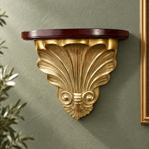 May include: A wall-mounted shelf featuring a dark brown, semi-circular wooden top surface supported by a decorative, gold-colored bracket. The bracket displays a prominent, carved fan-like shell motif with intricate, symmetrical scroll details.