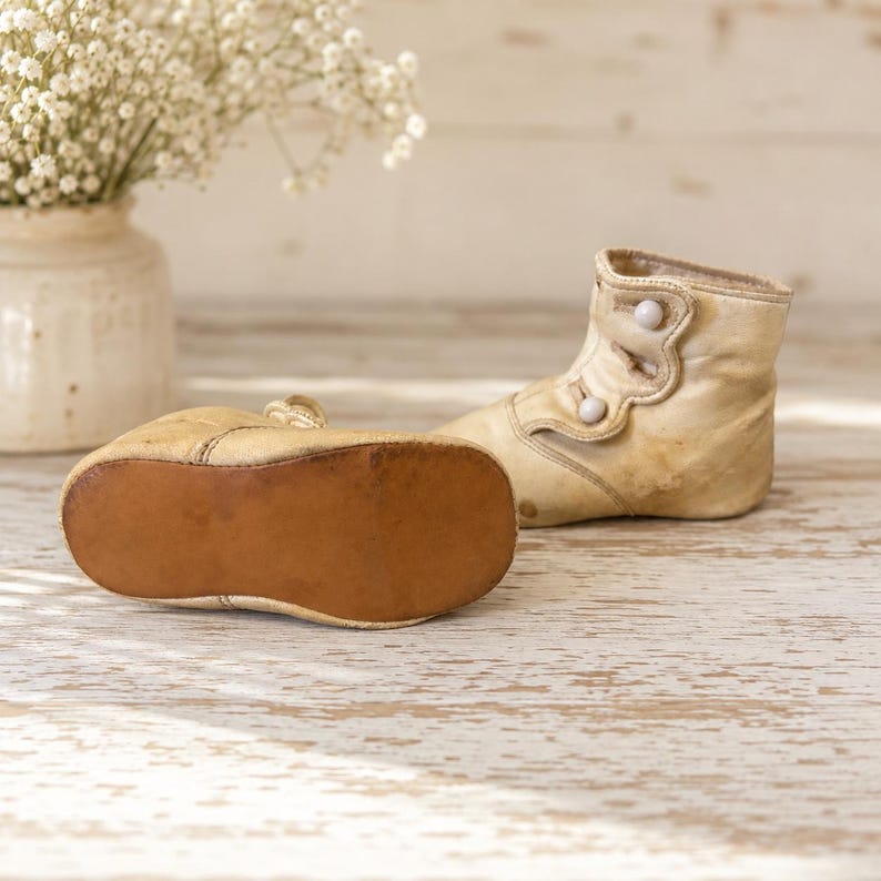 May include: A pair of vintage-style cream-colored leather baby boots featuring a high-top design with two small white buttons on the side. One boot is positioned upright, while the other is placed on its side to reveal a smooth, brown leather sole.