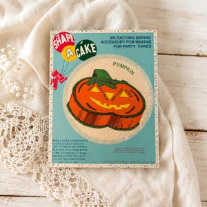 May include: A rectangular cardboard box for a pumpkin-shaped cake mold. The box features a teal background with a graphic of an orange jack-o'-lantern cake, red balloons, and text reading SHAPE A CAKE and PUMPKIN. It includes instructions for a 9-inch pan.
