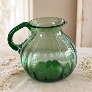May include: A translucent forest green glass pitcher with a rounded, bulbous body and vertical ribbed detailing. The vessel features a wide, open mouth and a curved handle extending from the side, crafted from smooth, polished green glass.