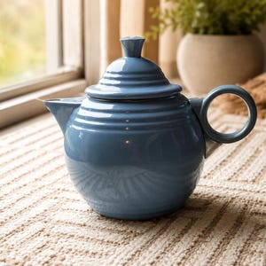 May include: A glossy, dusty blue ceramic teapot with a rounded body, a matching lid featuring a small knob handle, a curved spout, and a circular side handle. The exterior surface displays subtle horizontal ridges around the middle section.