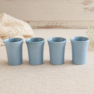 Könnte beinhalten: Ein Set aus vier kleinen, hellblauen Keramikbechern, die in einer horizontalen Reihe angeordnet sind. Jeder Becher hat eine glatte, glänzende Oberfläche, einen leicht ausgestellten Rand und einen konisch zulaufenden, zylindrischen Körper.