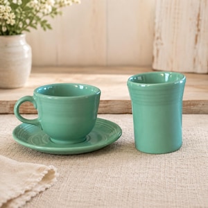 Può includere: Una tazza da tè in ceramica color turchese chiaro con piattino abbinato e una tazza cilindrica alta dello stesso colore. Entrambi i pezzi presentano una finitura liscia e lucida con sottili dettagli ad anello orizzontali sull'esterno.
