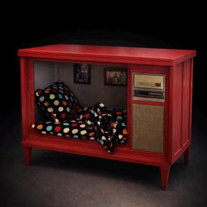 May include: A repurposed vintage red television cabinet transformed into a pet bed. The interior features a polka-dot pillow and blanket. The cabinet has a speaker and control panel on the side, and two framed pictures on the back wall.