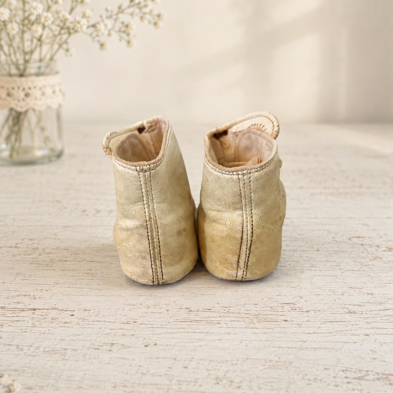 May include: A pair of metallic gold high-top booties featuring a smooth finish, visible vertical stitching along the back, and a small strap closure at the top. The footwear has a rounded toe design and a light-colored interior lining visible at the opening.