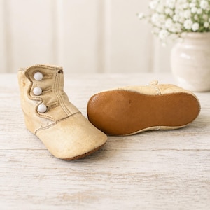 May include: A pair of cream-colored leather baby boots featuring a high-top design with three small white buttons along the side. One boot is standing upright to show the button detail, while the other is placed on its side to reveal the brown sole.