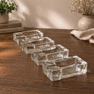 Op de afbeelding: Vier heldere glazen asbakken met een getextureerd oppervlak zijn gerangschikt op een houten tafel. Elke rechthoekige asbak heeft een klein, verhoogd gedeelte aan één kant. De asbakken zijn in een rij geplaatst, met een lichte hoek.