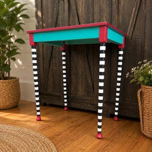 May include: A small rectangular table featuring a bright turquoise top frame with a solid magenta surface. The four legs are painted with alternating horizontal black and white stripes, each ending in a rounded magenta foot at the base.