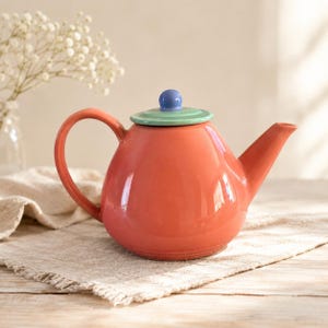 May include: A glossy coral-colored ceramic teapot featuring a rounded body, a curved handle, and a tapered spout. The lid is light green with a small, round, periwinkle blue knob handle centered on top, providing a distinct color contrast.
