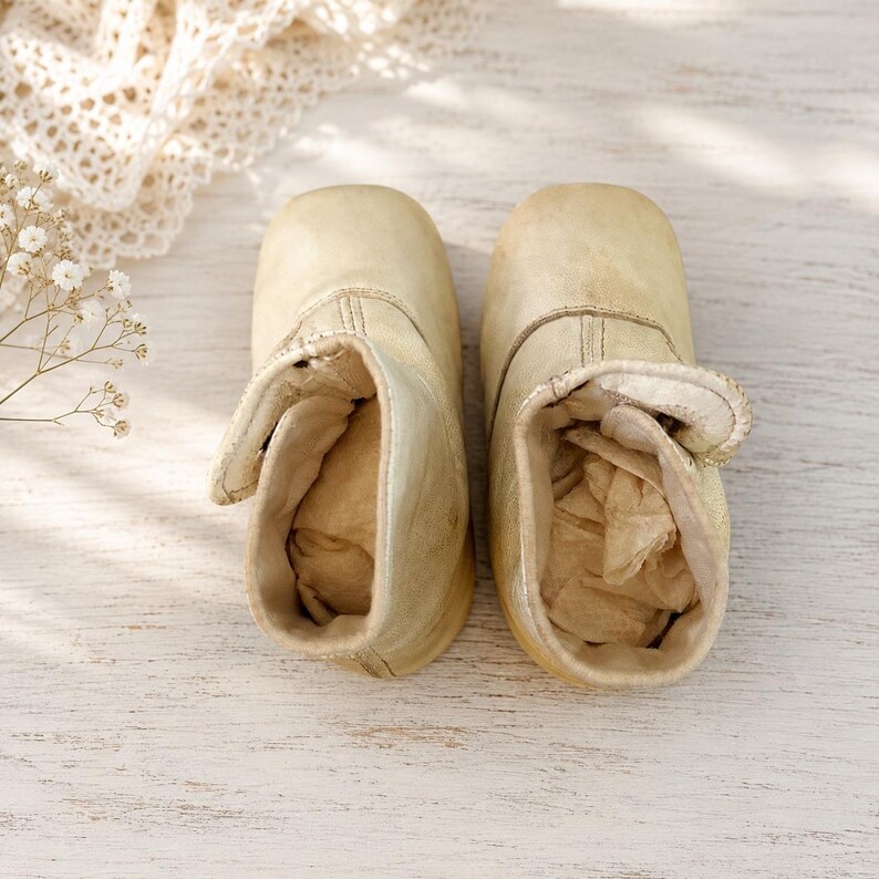 May include: A pair of cream-colored leather baby boots with a rounded toe, a high-top silhouette, and a side hook-and-loop closure. The interior of each boot is stuffed with light beige paper, and the exterior features visible stitching details.