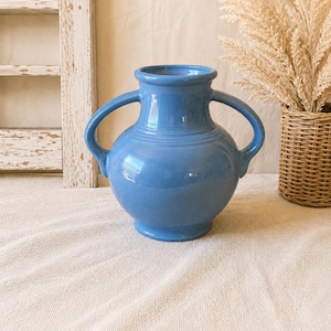 Könnte beinhalten: Eine glänzende, mittelblaue Keramikvase mit einem abgerundeten Körper, einem ausgestellten Rand und zwei symmetrischen, geschwungenen Henkeln an den Seiten. Die Oberfläche der Vase weist horizontale Rillendetails am Hals auf, die einen Kontrast bilden.