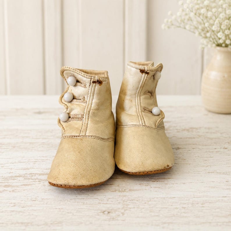 May include: A pair of off-white leather baby boots featuring a high-top design with three small, round, white buttons on the side of each boot. The boots have a rounded toe, a stitched seam across the vamp, and a visible texture on the leather surface.