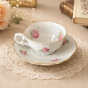 May include: A white porcelain teacup and saucer set featuring a delicate pink rose floral pattern and gold-trimmed scalloped edges. The teacup has a curved handle and a smooth finish, while the matching saucer displays a matching rose motif.