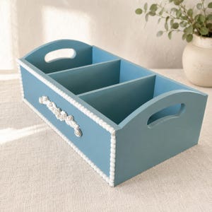 May include: A rectangular blue wooden storage caddy with three interior compartments and two side handles. The exterior edges are lined with small white beads, and a white floral relief ornament is centered on the front panel.