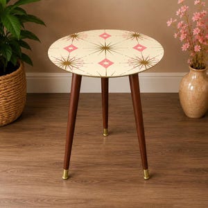 May include: A round side table with three tapered dark brown wooden legs featuring gold-toned metal feet. The tabletop has a cream-colored surface decorated with a repeating pattern of gold starbursts and small pink diamonds arranged in a grid.