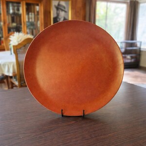 May include: A large, round, orange-brown plate displayed on a black metal stand. The plate has a smooth surface and is set on a dark brown wooden table. The plate is a warm, earthy tone.