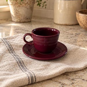 Puede incluir: Una taza de té de cerámica en color burdeos intenso con su plato a juego. La taza presenta un cuerpo redondeado con detalles acanalados horizontales en la parte superior y un asa curva. El plato tiene un acabado liso y brillante.