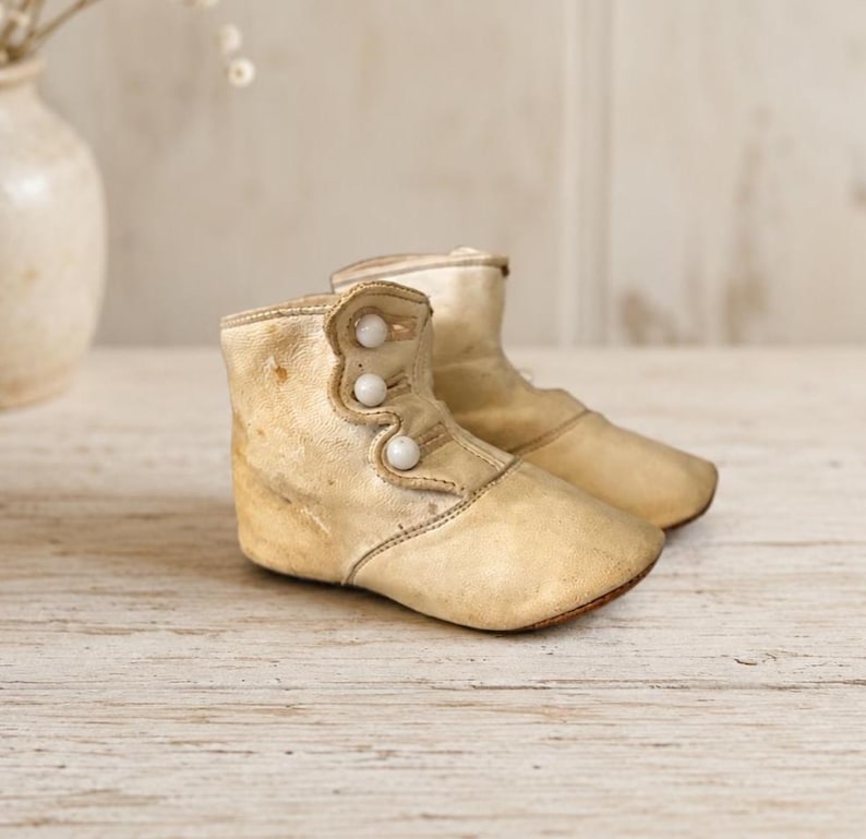 May include: A pair of vintage-style cream-colored leather baby boots featuring a high-top design with three small white spherical buttons along the side closure. The boots have a smooth, slightly worn texture and a rounded toe construction.