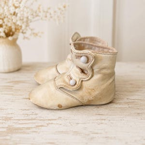 May include: A pair of off-white, ankle-high boots featuring a side-button closure with two small, round white buttons on each boot. The boots have a rounded toe, a structured collar, and visible signs of wear, including light brown discoloration.