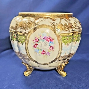 May include: A cream-colored porcelain bowl with a scalloped rim and three gold-painted feet. The exterior features ornate gold filigree, painted green garlands, and a central oval medallion displaying a colorful bouquet of pink, red, and white roses.