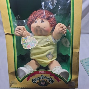 May include: A 1984 Cabbage Patch Kids doll with reddish-brown yarn hair and brown eyes, wearing a yellow romper with an elephant graphic and white shoes. The doll is inside its original yellow cardboard packaging with the text Cabbage Patch Kids.