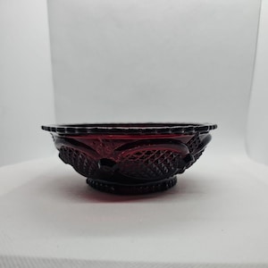 May include: A ruby red glass bowl with a decorative pattern. The bowl has a scalloped rim and a pedestal base. The exterior features a repeating design of diamond shapes and floral elements. The bowl is likely vintage or antique.