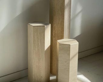 May include: Three beige, hexagonal pedestals of varying heights stand against a white wall. The pedestals have a natural, textured appearance, with visible grain patterns. The tallest pedestal is in the center, flanked by two shorter ones, creating a display.