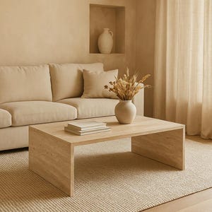 May include: A neutral-toned living room scene featuring a beige sofa, a rectangular wooden coffee table, and a beige rug. A vase with dried flowers sits on the table, with a stack of books. The room has a minimalist design with beige curtains and a wall niche.