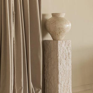 Könnte beinhalten: Eine beige, strukturierte Vase steht auf einem passenden Steinsockel. Die Vase hat einen abgerundeten Körper und einen schmalen Hals. Der Sockel ist rechteckig und hat eine raue, steinartige Textur. Beige Vorhänge links.