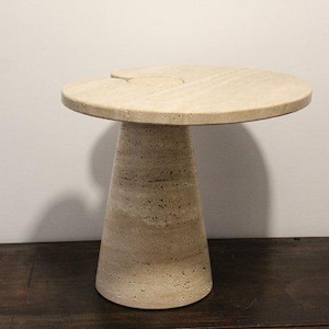 Può includere: Un tavolino in pietra beige con piano rotondo e base conica. Il tavolo ha un aspetto naturale e strutturato e una piccola tacca sul bordo del piano. Il tavolo misura circa 50 cm di diametro e 61 cm di altezza.