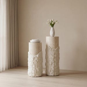 Könnte beinhalten: Zwei zylindrische Podeste mit einer strukturierten, hellfarbigen Steinbasis und einer glatten Oberseite. Ein Podest hält einen Stapel weißer Teller, das andere zeigt eine weiße Vase mit weißen Blumen. Die Podeste befinden sich in einem neutral getönten Raum.