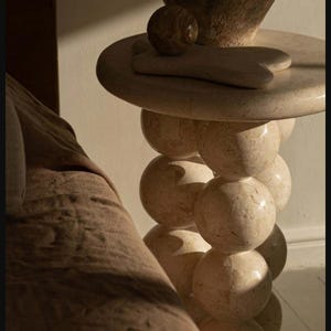 Può includere: Un tavolino in marmo beige con piano rotondo e base a sfere impilate. Un vaso decorativo, una piccola sfera e uno strumento per massaggio facciale poggiano sulla superficie del tavolo. Un mobile unico.