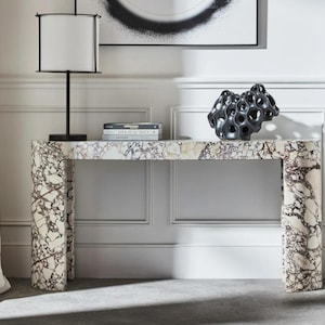 Può includere: Una consolle in marmo con un motivo bianco e bordeaux. Una lampada nera con paralume bianco e una scultura nera sono sopra. Il tavolo è sostenuto da due spessi gambe rettangolari.
