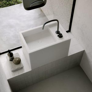 Op de afbeelding: Een moderne, minimalistische wastafel met een zwarte kraan en armaturen. De witte, rechthoekige wastafel staat op een verhoogd platform. Een handdoek, zeepdispenser en decoratief item staan op het platform.