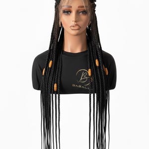 Peut inclure: Perruque tressée noire avec un devant en dentelle, présentée sur une tête de mannequin. La perruque présente de longues tresses individuelles ornées de perles en bois. Le mannequin porte un t-shirt noir avec un logo.