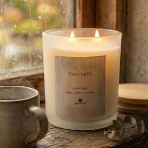 Op de afbeelding: Een brandende sojawas kaars in een matglazen pot. De kaars heeft een houten label met de tekst "FIRST RAIN". De kaars is handgegoten en weegt 150 gram. Een keramische mok en een houten onderzetter staan op de achtergrond.