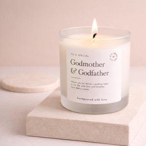 Op de afbeelding: Een brandende witte kaars in een matglazen pot. Het etiket luidt "TO A SPECIAL Godmother & Godfather" met een bericht eronder. De kaars staat op een lichtgekleurd, vierkant blok. Onderaan staat "handpoured with love".