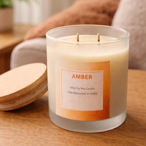 May include: A frosted glass candle with two wicks and a wooden lid. The candle has a label that reads "AMBER" and "150g Soy Wax Candle Handpoured in India". The candle is on a wooden surface.