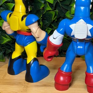 May include: Two action figures, Wolverine and Captain America, are displayed from the back. Wolverine is yellow and blue, while Captain America is blue, red, and white. The figures are on a wooden surface with a green leafy background.