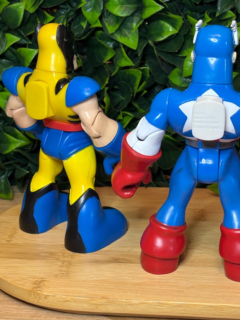 May include: Two action figures, Wolverine and Captain America, are displayed from the back. Wolverine is yellow and blue, while Captain America is blue, red, and white. The figures are on a wooden surface with a green leafy background.