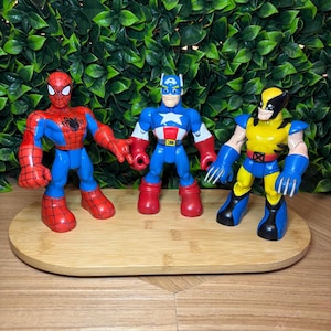May include: Three superhero action figures: Spider-Man, Captain America, and Wolverine. Spider-Man is red and blue, Captain America is blue, red, and white, and Wolverine is yellow and blue. They stand on a wooden tray.