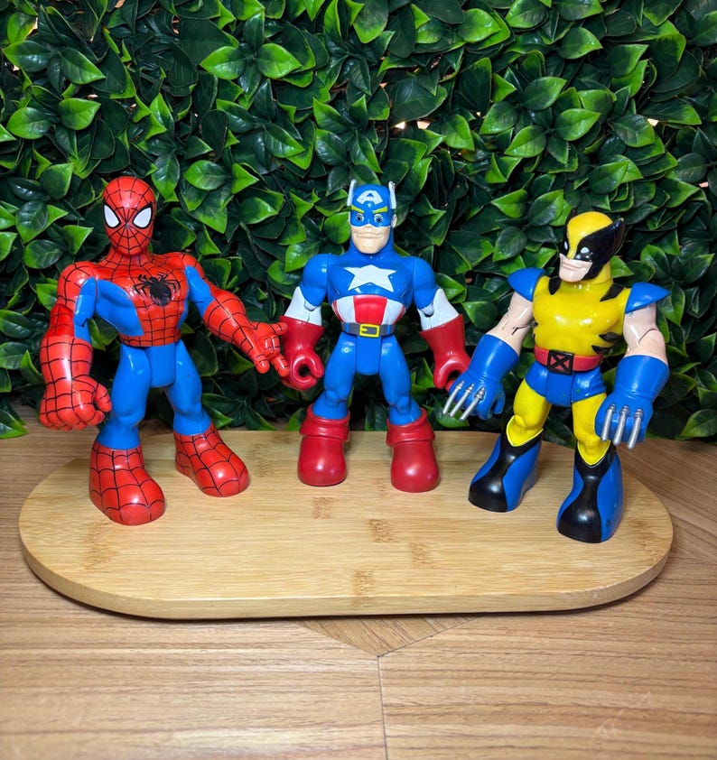 May include: Three superhero action figures: Spider-Man, Captain America, and Wolverine. Spider-Man is red and blue, Captain America is blue, red, and white, and Wolverine is yellow and blue. They stand on a wooden tray.