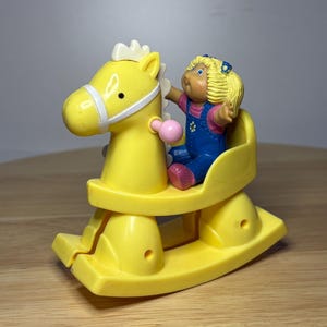 May include: A yellow plastic rocking horse toy with a white bridle and a small doll figure wearing blue overalls and a pink shirt. The doll has blonde hair and is seated on the horse. The toy is on a wooden surface.