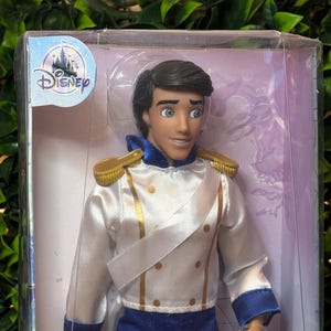 May include: A Disney Prince doll in a clear plastic display case. The doll wears a white satin jacket with gold trim and blue pants. The Disney logo is visible on the packaging.