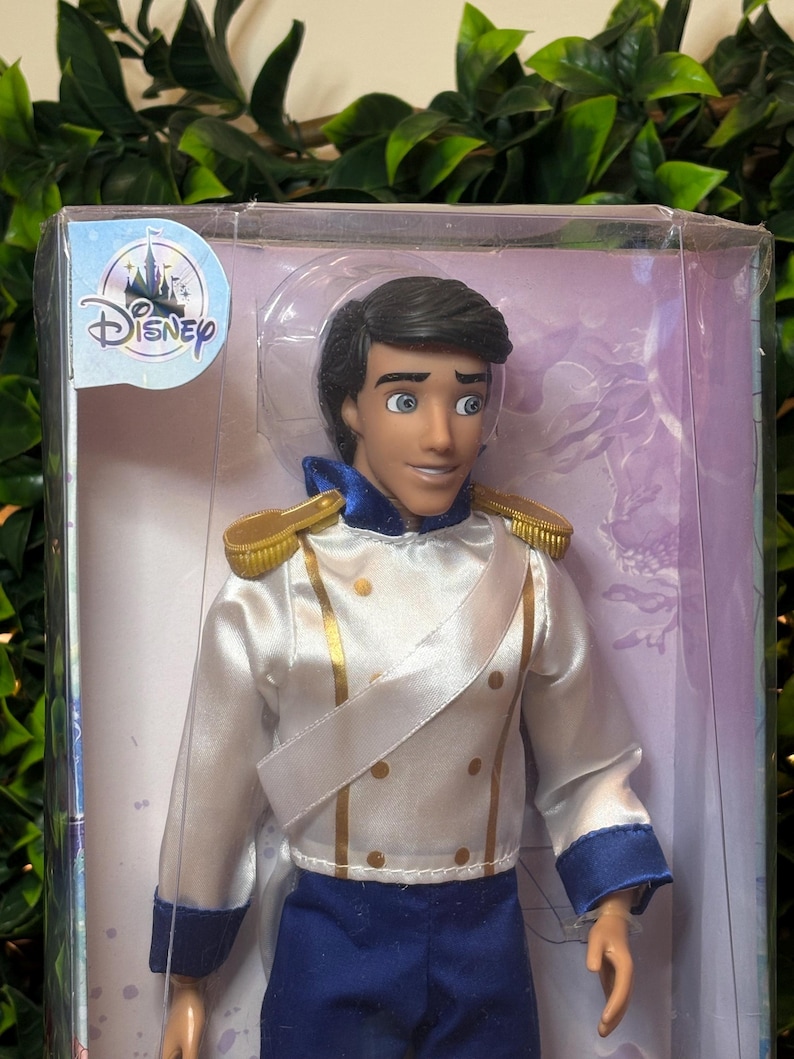 May include: A Disney Prince doll in a clear plastic display case. The doll wears a white satin jacket with gold trim and blue pants. The Disney logo is visible on the packaging.