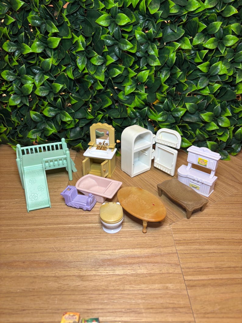 May include: A collection of miniature dollhouse furniture including a light green crib with a slide, a yellow vanity with a mirror, a white refrigerator, a pink bathtub, a purple toy train, a toilet, an oval wooden table, and a white kitchen unit.