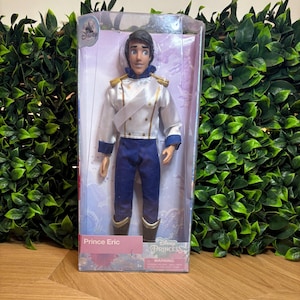 May include: A Disney Princess Prince Eric doll in a clear plastic box. The doll wears a white jacket with gold trim, blue pants, and black boots. The box has the Disney Princess logo and the name "Prince Eric".