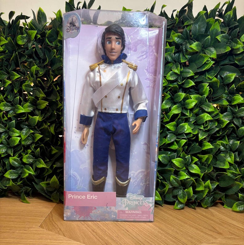 May include: A Disney Princess Prince Eric doll in a clear plastic box. The doll wears a white jacket with gold trim, blue pants, and black boots. The box has the Disney Princess logo and the name "Prince Eric".