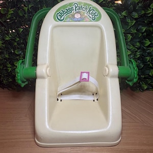 Peut inclure: Un siège porte-poupée Cabbage Patch Kids en plastique crème doté d'une poignée réglable verte avec des boutons latéraux en forme de fleur. Le siège comprend une sangle de sécurité blanche avec une boucle rose et le logo Cabbage Patch Kids.