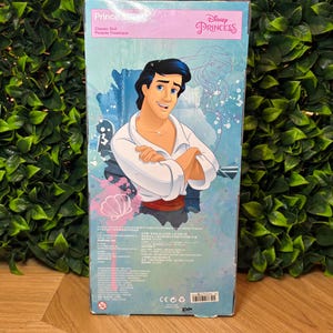 May include: A Disney Princess Prince doll box featuring a cartoon illustration of a man with black hair, wearing a white shirt and red sash. The box has the words "Prince" and "Classic Doll" printed on it.